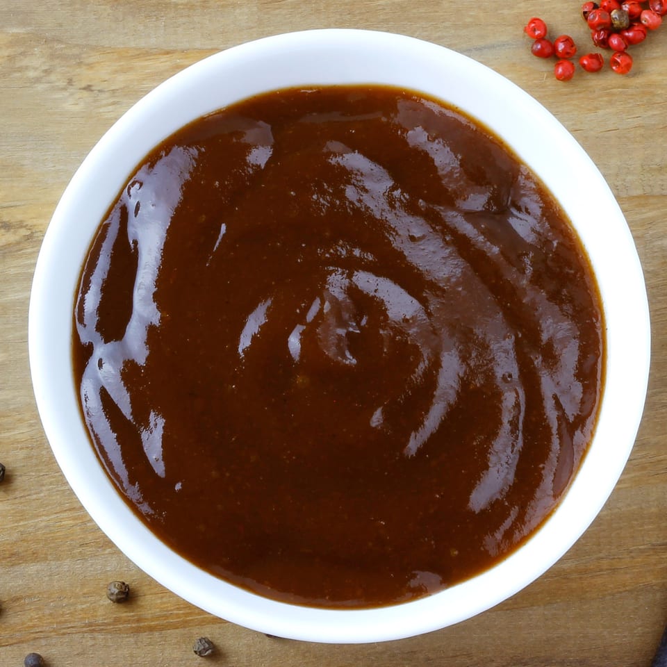 A white bowl filled with rich, dark brown barbecue sauce, on a wooden surface.