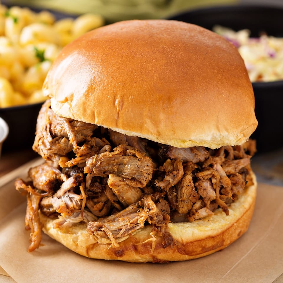 Close-up of a Fisher's BBQ & Moore pulled pork sandwich on a bun with sides in the background.